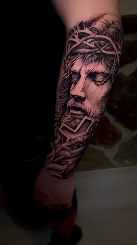 Rostro de Cristo con corona de espinas y cruz en el antebrazo, realismo en negro y gris